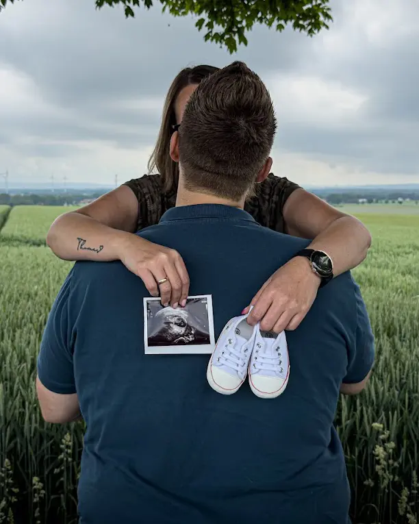 Zwei Menschen von hinten in einem Feld, in den Händen ein Ultraschallbild und Babyschuhe